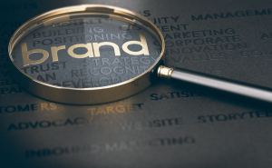 Magnifying glass highlighting the word “brand” on a dark background filled with related branding terms such as positioning, brand strategy, trust, recognition, and marketing.